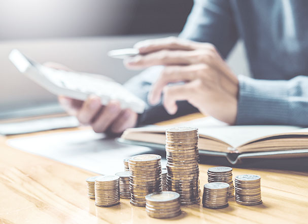 Stacks of coins of different sizes on a table. In the background, a person using a calculator