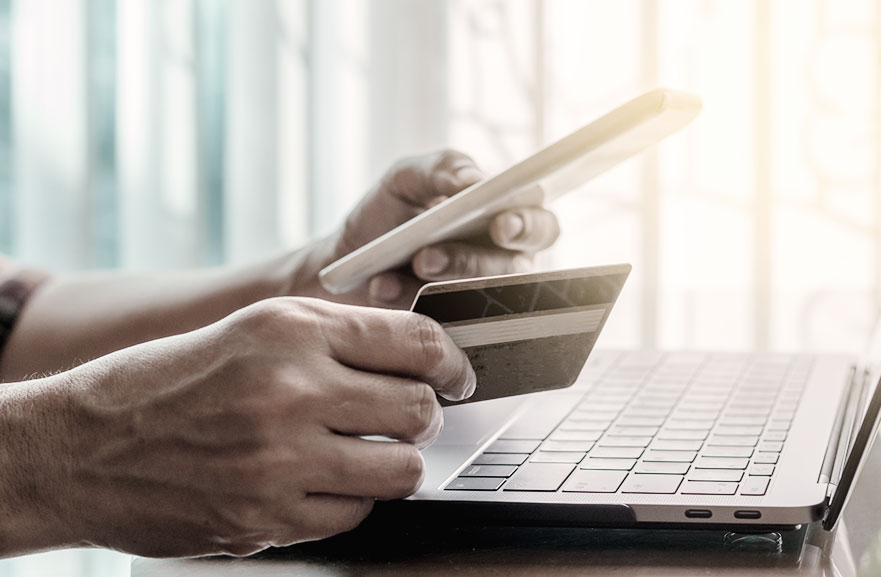 Person holding a credit card in one hand while typing on a cell phone with the other. In the background, a silver notebook is positioned above a table