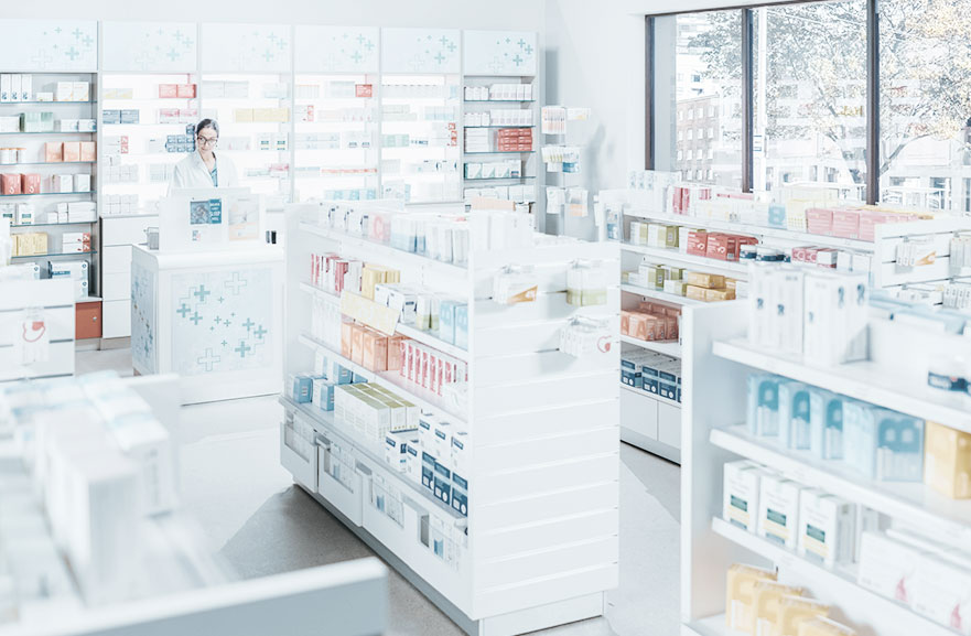 Visão interna de estabelecimento farmacéutico, com gondolas brancas e prateleiras com medicamentos.  