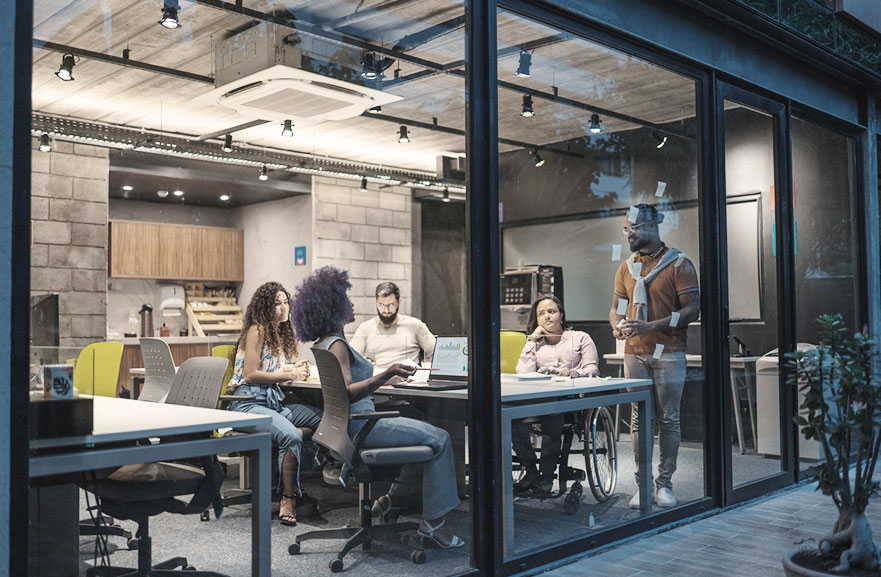 Grupo de cinco pessoas em reunião dentro de uma sala com paredes de vidro