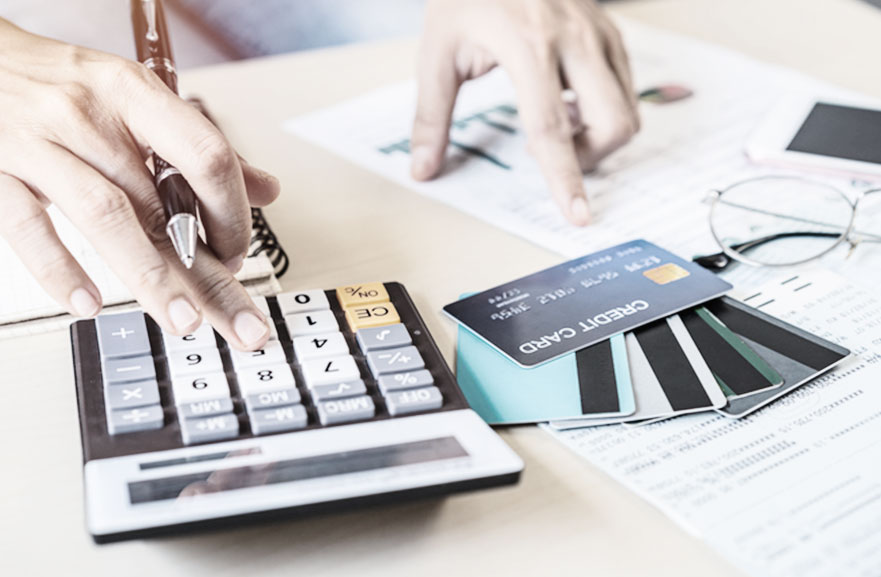 A person doing math on a calculator. Next to it, on the table, several credit cards and sheets of paper