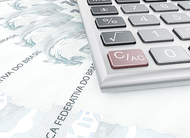 Calculadora cinza, com botões em cinza, preto e vermelho, posicionada acima de cédulas de reais