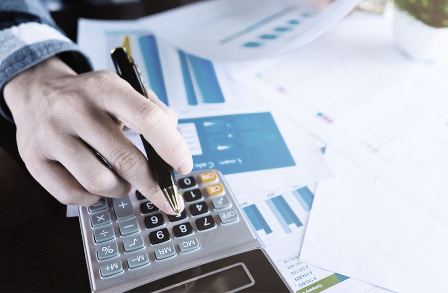 Pessoa utilizando calculadora cinza com teclas em preto e cinza. Ao lado, estão distribuidas várias folhas de papel sulfite branco com gráficos impressos