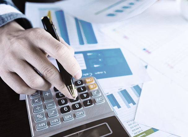 Pessoa utilizando calculadora cinza com teclas em preto e cinza. Ao lado, estão distribuidas várias folhas de papel sulfite branco com gráficos impressos