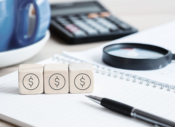 Blocos de madeira 3d posiconados acima de caderno com folhas brancas. Mais ao fundo, calculadora preta com teclas brancas e vermelhas e lupa na cor preta.