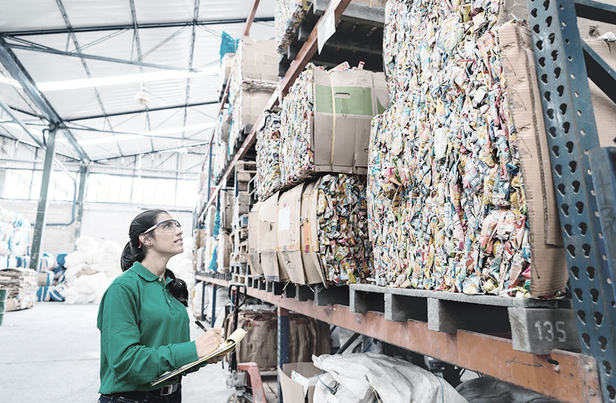 Mulher usando camiseta verde, óculos de proteção e cabelo preso com rabo de cavalo, segurando prancheta e olhando para estoque de materiais em uma fábrica