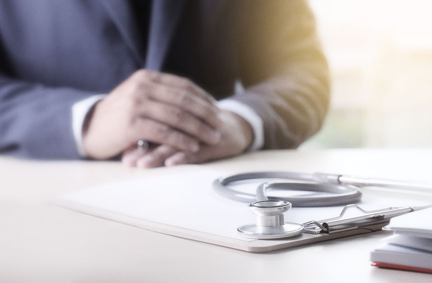 Prancheta beje com folha sulfite branca e estetoscópio acima, posicionados em cima de uma mesa.