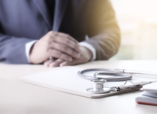 Prancheta beje com folha sulfite branca e estetoscópio acima, posicionados em cima de uma mesa.