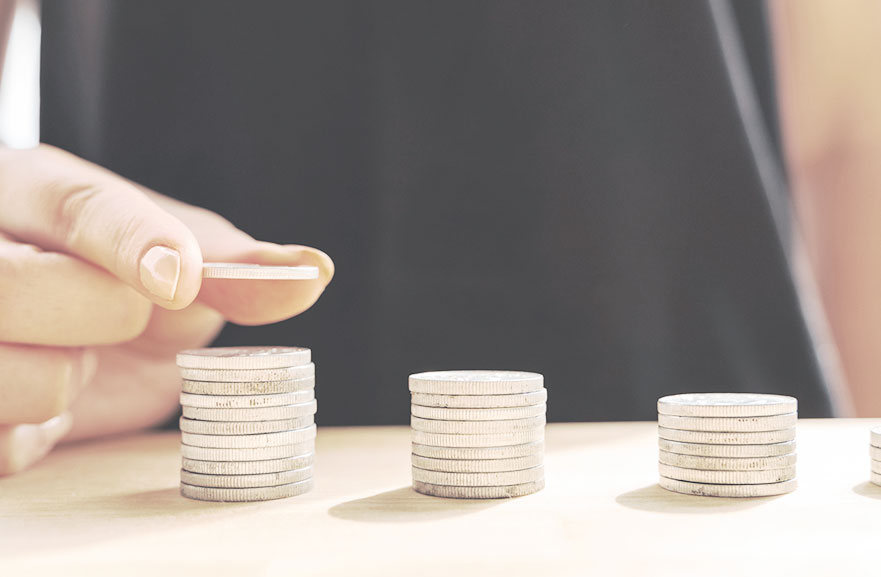 Três pilhas de moeda, de diferentes tamanhos. Na pilha da esquerda, uma pessoa colocando mais uma moeda