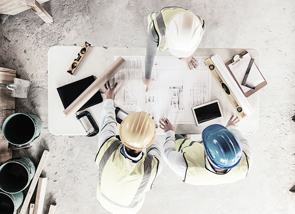 Visão superior de três pessoas em um ambiente em obra. Eles estão usando capacetes, equipamentos de segurança e ao redor de uma mesa.