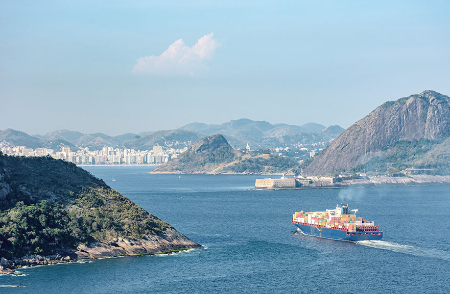 Vista superior do oceano, com navio cargueiro atravessando. Mais ao fundo, está a cidade do Rio de Janeiro