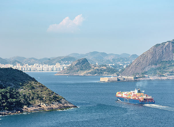 Vista superior do oceano, com navio cargueiro atravessando. Mais ao fundo, está a cidade do Rio de Janeiro