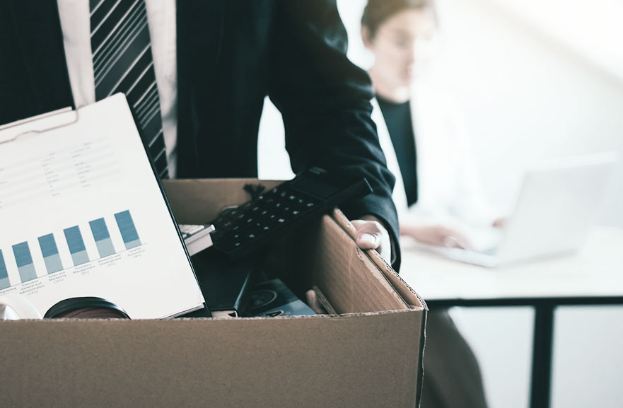 Executivo usando camisa branca, gravata listrada e paletó preto, segurando uma caixa de papelão com materiais de escritório dentro.