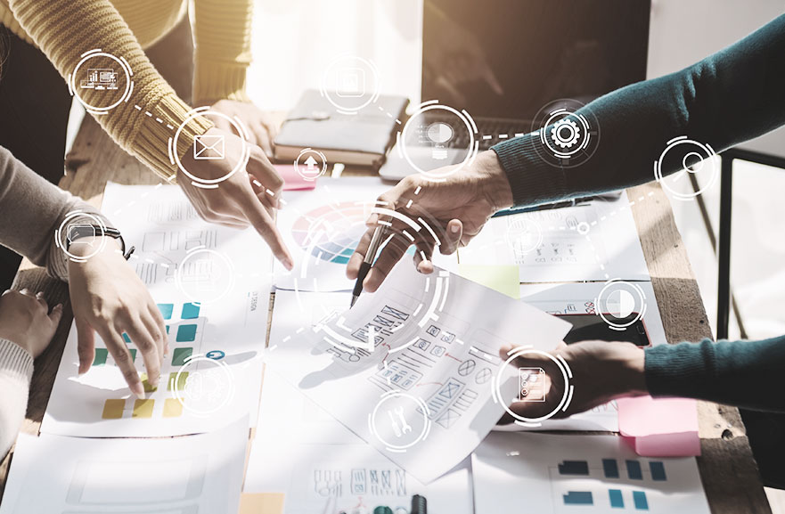 Três pessoas ao redor de uma mesa com vários papeis sulfites acima dela. Nos papeis, estão impressos infográficos e textos